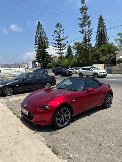 In the city of Mesogi, Cyprus, on a bright, sunny day, a red Mazda MX-5 convertible is parked on the right side of the frame, angled towards the left. The car's black soft top is up, and its license plate reads "NZV-451". To the left of the Mazda, a dark gray BMW sedan is parked, facing right. Further to the left, a white van with the branding "euroblinds" is parked. In the background, several other vehicles are visible on a road, including a white Nissan Qashqai and a black Jeep. The environment is a street scene with a sidewalk on the left and a paved road on the right. Tall, slender evergreen trees line the background, and a blue sky with scattered white clouds is overhead. A building with signs is visible behind the vehicles on the road.