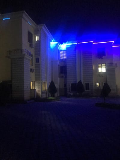 The image depicts a multi-story building complex at night in Ushapa, Nigeria. The building features light-colored exterior walls, multiple windows with some illuminated interior spaces, and prominent vertical architectural elements, including pillars and sections with balustrades. Extensive blue LED strip lighting is installed along the roofline and architectural cornices of the structure, casting a distinct blue glow that illuminates the upper portions of the building and reflects faintly on the ground. Several air conditioning units are mounted on the exterior walls. In front of the building is a dark, paved surface, consistent with interlocking pavers. Multiple small, conical evergreen trees are interspersed within landscaped sections directly adjacent to the building's base. The sky is dark, indicating nighttime conditions. No individuals or discernible actions are present within the scene.