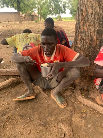 The image shows a young person sitting on the ground, leaning against a tree trunk and eating from a metal bowl with a spoon. The person is wearing an orange t-shirt and grey shorts. Their skin is dark and appears dusty. In the background, other people are partially visible, one wearing a yellow t-shirt with text, and another in a colorful patterned fabric. The ground is sandy and dirt, with exposed tree roots. The setting appears to be outdoors, likely in a village, with some vegetation and rudimentary structures visible in the distance. The lighting suggests it could be daytime, possibly overcast or late afternoon. The overall scene conveys a sense of everyday life, with the focus on the individual eating. The visible text on the yellow t-shirt reads "BOTT NL".