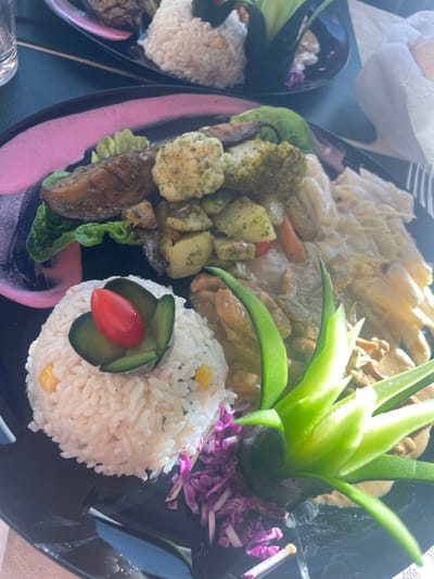 The image presents a close-up, high-angle view of two prepared meals served on black plates, positioned on a dark table surface within a dining establishment in Lkheng, Morocco.

The primary foreground plate, in sharp focus, displays a variety of dishes. A dome of white rice, interspersed with corn kernels and green herbs, is crowned by a red cherry tomato flanked by two curved green cucumber slices. To the left, a piece of seared fish rests on a bed of green lettuce. Adjacent to the fish, a medley of cooked vegetables including cauliflower florets, broccoli florets, cubed potatoes, and carrot slices is coated in a light green herb sauce. To the right, penne pasta is served in a creamy, light-colored sauce, accompanied by pieces of chicken or similar meat in a yellowish-green sauce. A vibrant pink sauce is artfully swirled along the left rim of the plate. At the bottom right, a decorative carving from a green vegetable, possibly cucumber, resembles a stylized flower. A small pile of shredded red cabbage is situated below the rice.

The secondary plate, visible in the blurred background, features a similar presentation, including a mound of rice and elements of a pink sauce on its rim. A clear drinking glass is partially visible on the far left, and a white napkin or bread basket is partially visible on the far right. No individuals are present in the scene.