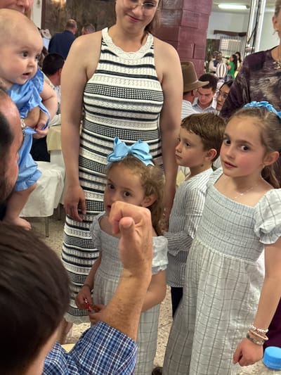 The image captures an indoor scene with multiple individuals. The foreground features a baby wearing blue, held by an adult, and a woman in a black and white striped dress. Three children are also visible: two girls, one with a blue bow in her hair, and a boy, all in striped or patterned clothing. One of the girls has bracelets on her right wrist. An adult is also partially visible, wearing a blue plaid shirt.
Background details include other people, some wearing hats, and what appear to be rows of objects, possibly boxes or containers, stacked along a wall. Fluorescent lighting is visible on the ceiling. The image appears to have been taken at an event. Without additional details, specific events or locations in Chipilo are indeterminable.