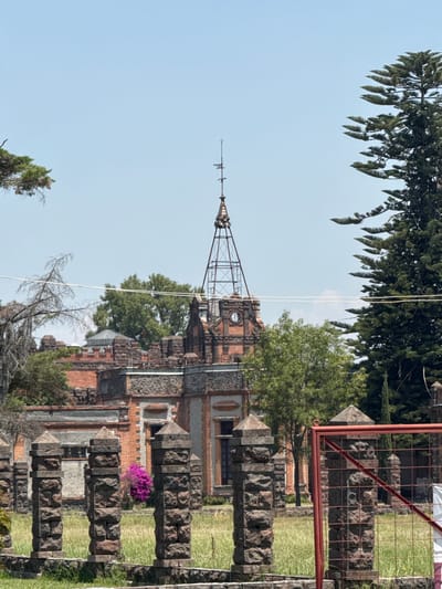 The image depicts a tall building with a spire, possibly a church or historical structure, in Santa Ana Portales, Mexico. The building's architecture features brick and stone elements. In the foreground, a stone wall or barrier is visible, with grass and purple flowers growing nearby. Overhead, trees partially obscure the sky. A metal fence is seen on the right side of the frame.