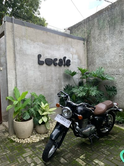 The image displays an outdoor courtyard entrance of a business named "Locale" in Kota Batu, Indonesia. A textured concrete wall serves as the primary backdrop, featuring the dark, stylized "Locale" sign, which incorporates a coffee cup design into the letter "o." In the foreground, a black, classic-style motorcycle with a ribbed brown leather seat is parked on a kickstand, facing left. Its front license plate reads "L 2362 IR," indicating registration in Surabaya, with "03-29" below. Abundant green vegetation is present, including three potted plants on white decorative stones to the left of the motorcycle, and a large-leafed plant situated to its right, against the wall. The ground is paved with rectangular bricks, many of which are covered in green moss. A second, moss-covered concrete wall forms a corner on the right side of the scene. A wooden door frame is partially visible on the far left. The sky is overcast. No individuals or active interactions are present in the scene.