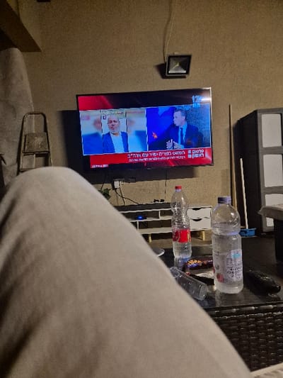 The image depicts an indoor scene in Rehovot, Israel, from a first-person perspective, focused on a television screen and a coffee table. The lower left and center foreground are occupied by the upper thigh and knee of a person wearing light-colored, possibly beige, pants.
On a dark, possibly woven, coffee table in the midground, several objects are present: a clear plastic bottle with a red label (likely water), a clear plastic bottle with a blue and white label displaying Hebrew text (partially full), and an empty, clear plastic bottle lying horizontally. A small, dark, rectangular package, possibly a snack, is visible, along with a black remote control.
A flat-screen television is mounted on a light-colored, possibly beige, wall in the background. The television displays a split-screen news broadcast from Channel 12, as indicated by the numeral "12" in the upper right. On the left side of the screen, a man with light hair and a grey suit is shown. On the right, a man in a dark suit is visible, gesturing with his hands. Hebrew text, likely a news ticker or headline, is displayed in a red banner across the bottom of the screen. Below the television, a white, multi-tiered stand holds an electronic device with a blue indicator light on its lower shelf.
To the left of the television, a folded metal object, resembling a small ladder or chair, leans against the wall. Above and to the right of the television, a wall-mounted square light fixture is visible, with an electrical wire extending upwards. To the far right, a dark, modular storage unit or cabinet with distinct square compartments is partially visible, with a long, thin, light-colored pole leaning against it. The overall lighting suggests an indoor evening or dimly lit environment. No direct actions beyond viewing the television are captured, and no direct interactions between individuals are depicted.