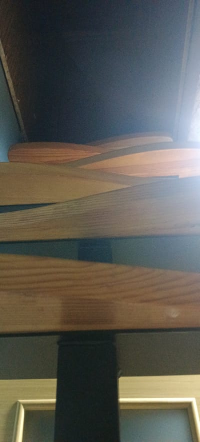 The image displays a close-up, angled view of wooden slats, likely part of a bed frame or shelving unit. The slats are light brown with visible wood grain. Some slats are positioned horizontally, while others are angled, creating overlapping layers. The background is dark, with a hint of lighter wood at the top. In the lower portion of the image, a black, vertical object is visible, suggesting a supporting structure. Below this, a light-colored frame with a beveled edge is partially visible, possibly part of a furniture item or a doorway. The lighting is uneven, with a bright glare on the right side of the image, indicating a light source from that direction. The scene appears to be indoors, in a domestic setting, possibly within a residential area of Хацежынскі сельскі Савет, Belarus. There are no people or animals present in the image.
