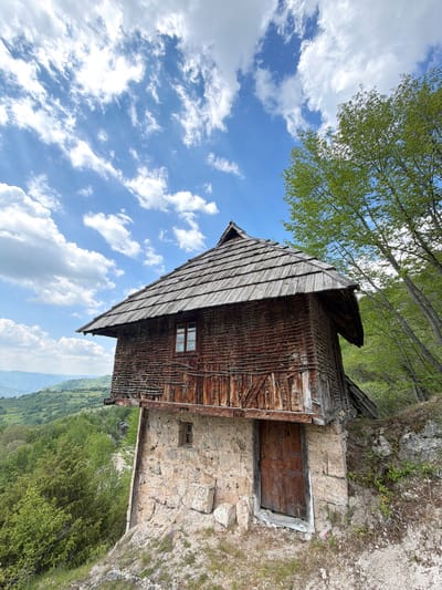 A landscape photograph depicts a traditional wooden structure, likely a rural home or outbuilding, set against a backdrop of a partly cloudy sky. The wooden building features a steeply pitched roof covered with wooden shingles, a small window, and rustic exterior walls constructed from logs. A section of the building appears to be constructed from stone. A wooden door is attached to the stone section of the building. The environment includes a lush green landscape, possibly a valley or hillside, and vegetation, including trees with light green foliage. The sky is blue with white clouds. The image is likely taken in a rural area of Prijepolje Municipality, Serbia.