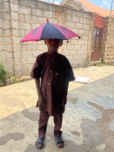 A male child, approximately 5-10 years old, stands centered in an outdoor setting in Idiroko, Nigeria, facing directly towards the viewer. He is dressed in a maroon two-piece garment consisting of a short-sleeved tunic with gold-toned embroidered patterns on the chest and matching trousers, along with dark grey slide sandals on his feet. A small, open umbrella, featuring alternating black and magenta-pink panels with decorative tassels along its rim and a red finial, is balanced on his head. The background primarily consists of a grey concrete block wall. To the child's right, a reddish-brown metal gate with vertical bars is integrated into the wall, beyond which a structure with a reddish corrugated metal roof is visible against a bright sky. Sparse green vegetation grows along the base of the concrete wall to the child's left. The ground is a light grey concrete slab exhibiting areas of wear and discoloration. A white rectangular object lies flat on the concrete surface to the child's right. The scene is illuminated by natural daylight.