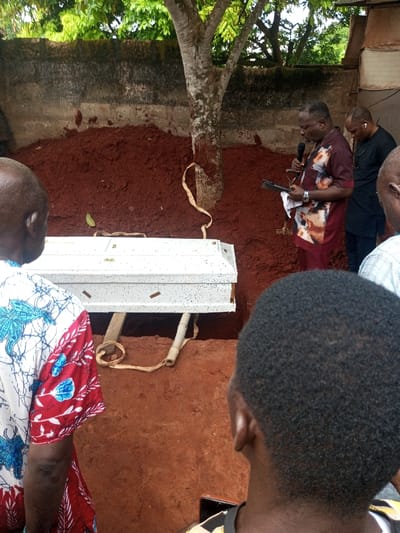 The scene depicts an outdoor burial ceremony in Oko Town, Nigeria. A white, speckled casket with dark flecks rests horizontally over an open grave, supported by two wooden beams and a cylindrical object, with beige fabric straps visible beneath it, presumably for lowering. A substantial mound of reddish-brown excavated soil is piled behind and to the right of the grave.
To the right of the casket, a male individual stands, wearing a short-sleeved collared shirt with maroon and abstract brown/white patterns, and maroon trousers. He holds a black microphone in his left hand and a dark tablet or book in his right, with a white cloth draped over his right forearm, appearing to be conducting a service or giving an address. Behind and slightly to his right, a second male individual, wearing a dark tunic and earphones, is partially visible.
In the immediate foreground, two individuals are seen from behind, observing the scene. On the left, a person is wearing a white shirt with prominent blue and red patterns. On the right, a person with short, dark, kinky hair is visible from the back of their head and upper shoulder.
In the background, behind the soil mound, a tree with a light-colored, mottled trunk stands. Behind the tree and soil is a weathered concrete wall. Above the wall, green foliage from other trees is visible. The ground throughout the visible area is reddish-brown soil.