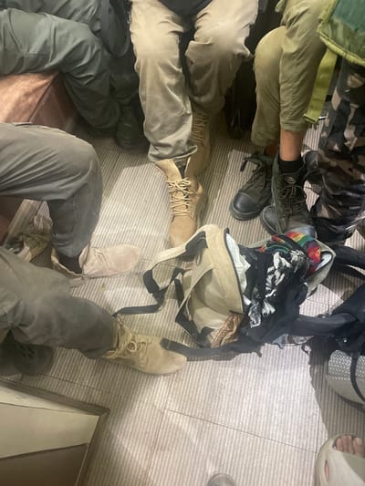 The image shows a close-up, overhead view of multiple people seated on a patterned floor. Five sets of legs and feet are visible, dressed in various pants and boots. The pants include camouflage, green, khaki, and gray materials. The footwear consists of several pairs of boots, including tan, gray, and dark green styles. There is also a bare foot wearing a sandal.
Between the legs, there are backpacks and other bags, including one open bag with what appears to be clothing inside. The setting is indoors with a tiled or carpeted floor.
The specific location of Mira-Bhayander, India is not evident from the details in the image. Therefore, I cannot pinpoint an exact location within the city.