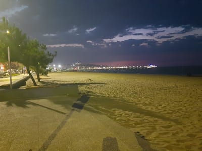 The image depicts a nighttime scene on a beach in Atakum, Turkey. A sandy beach stretches across the foreground and midground, with footprints and shadows cast by trees and lighting fixtures visible on the sand. A paved walkway runs along the left side of the image, bordering the beach. Several trees with green foliage are present on the left side of the walkway. In the distance, a coastline is illuminated by numerous lights, suggesting a populated area. The sky is dark with dramatic, scattered clouds, indicating it is either late evening or early morning. The overall atmosphere is serene, with the lights from the distant city reflecting on the water.