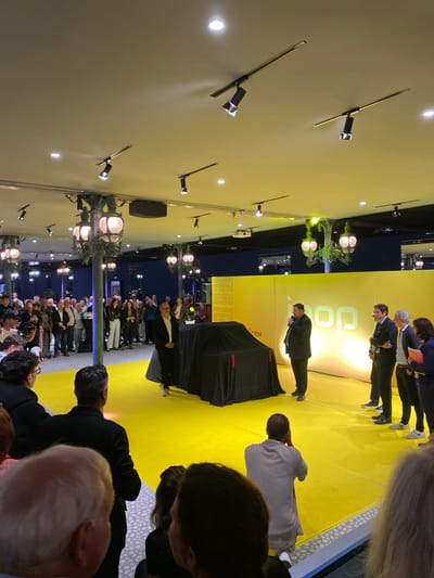 The rotated image captures an indoor event, likely a presentation or product launch, in Mulhouse, France. The scene features a brightly lit stage with a yellow floor and backdrop, contrasting with a darker audience area.

On stage, a man in a dark suit stands behind a black-draped podium, addressing the audience. He appears to be speaking into a microphone. Behind him, a large yellow banner displays the stylized logo "POP" and "EDF" (Électricité de France, a French electric utility company). Below these, text visible on the banner reads "PREMIERE MONDIALE" (World Premiere), "25 AVRIL 2023", and "MULHOUSE," indicating the event's nature, date, and location.

Further back on the yellow stage, three other individuals are present: two men in suits and a woman in dark attire. One man, wearing a white shirt and black jacket, is seen bowing towards the audience.

In the foreground and extending into the mid-ground, numerous audience members are gathered, watching the stage. Many are partially obscured, with several heads visible in the immediate foreground, suggesting the viewer is among the crowd. One person in a light grey outfit and another in a dark jacket are seen from behind, facing the stage.

The venue features a light-colored ceiling equipped with multiple spotlights providing bright illumination. Decorative lampposts with glowing globe lights are spaced throughout the event space, adding to the indoor ambiance. Audio speakers are also visible, mounted near the ceiling. The overall impression is a formal public event with speakers and an attentive audience.