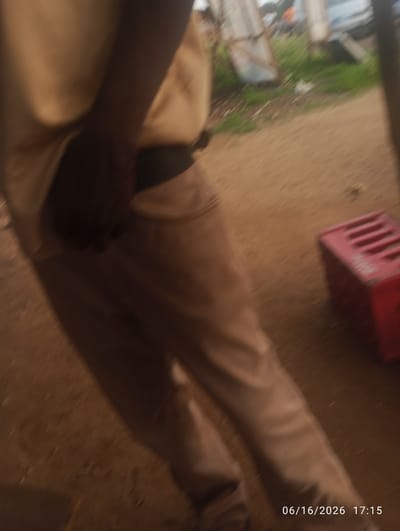 A close-up, slightly blurred, medium shot captures the lower body and arm of a person wearing a beige shirt and light brown trousers. The person's left arm is bent and appears to be holding something. The background is a mixture of dirt, grass, and some out-of-focus structures, suggesting an outdoor urban or semi-urban environment. To the right, a bright red crate with a slotted design is visible. The lower right corner of the image displays a timestamp: "06/16/2026 17:15". The scene appears to be in the city of Dokan Tofa, Nigeria.