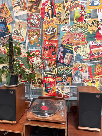 This image depicts a wall covered in vintage-style posters, reminiscent of a retro diner or an old movie theater. The posters feature various themes, including movie nights, music festivals, advertisements for beer and coffee, and depictions of people in period clothing. In the foreground, a wooden stand holds two audio speakers, a turntable with a vinyl record, and a plant in a black pot. The overall aesthetic is a blend of retro and contemporary. The location in Tam Kỳ, Vietnam, is not discernible from the provided image.