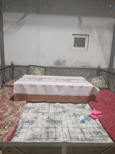 The image depicts a low-angle, close-up view of a covered bench or low platform, likely outdoors and possibly in a courtyard setting within a residential area of Социализм, Kyrgyzstan. The platform is covered with three distinct rugs, one red with a floral pattern, another with a muted, abstract pattern, and a third with a dark patterned border.
Dominating the center is a table covered with a white tablecloth edged with a brown border and a decorative band. Two patterned cushions, one green with an intricate design and another with a similar but less visible pattern, are placed at either end of the bench, leaning against a decorative metal railing.
In the background, a plain white wall with some visible staining and discoloration is present. A small, square window with a patterned glass pane is visible in the upper right portion of the wall. The overall lighting suggests it is either dusk or nighttime, with shadows cast on the wall from an unseen light source. A small, blue and white stuffed toy and a pink, folded piece of paper or origami are visible on the rug in the foreground.