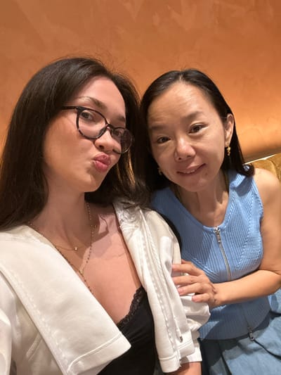 Two East Asian adult females are depicted in a close-up indoor portrait. The woman on the left, with long dark hair and dark-rimmed glasses, wears a white open collared jacket over a black lace-trimmed top, accessorized with layered gold necklaces. She puckers her lips in a 'duck face' expression while looking directly towards the camera. Her right hand rests on the upper left arm of the other woman. The woman on the right, with dark hair parted down the middle, wears a light blue sleeveless ribbed top with a silver front zipper and gold earrings. She smiles gently, also looking at the camera, with her left hand resting on the left woman's shoulder. The background is a plain, warm orange-brown wall, indicative of an interior setting. The specific location within Paradise, United States, is not discernible from the visual information provided.