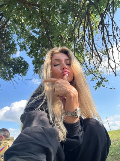 This is a portrait of a woman with long blonde hair, taken outdoors during the daytime. She is wearing a black hooded garment and a silver watch on her left wrist. Her eyes are closed, and she is puckering her lips while touching them with her index finger, creating a playful or suggestive expression. Her blonde hair is spread out, some of it falling onto her face and shoulder.
Behind her, the scene is dominated by a clear blue sky with scattered white clouds. Lush green trees with intricate branches frame the upper left portion of the image. In the background, a field is visible, and a utility pole with wires stands against the sky, suggesting a rural or semi-rural setting.
The overall mood is casual and lighthearted, captured in what appears to be a sunny day, possibly late morning or early afternoon, given the bright light. The angle of the shot, combined with the subject's pose, suggests a selfie. There is no visible text in the image. The location context provided is "Фельдмаршальский, Russia."
