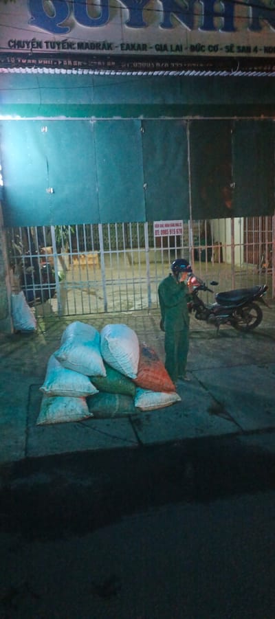 At night, a person in a green uniform stands next to a pile of sacks. The sacks are of various colors, including white, green, and red, and appear to be filled with goods. A motorcycle is parked behind the person. In the background, there is a metal gate and a building with a sign that reads "CHUYỀN TUYẾN MADRAK - EAKAR - GIA LAI - ĐỨC CƠ - SẼ SAN 4 - KONT". Another smaller sign is visible with the text "Đt: 0985 815 678". This scene is located in Xuân Long, Vietnam.