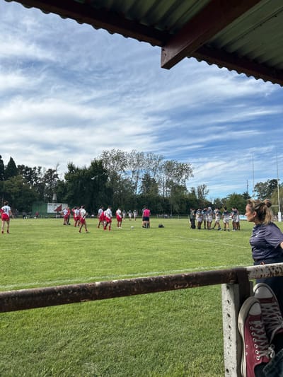 The image depicts a rugby match in Moreno, Argentina. Two teams, one in white and red and the other in white and blue, are lined up facing each other on a green field marked with white lines. Trees are visible in the background, along with a building. A woman is visible, standing over the scene. The image appears to be taken from under a corrugated iron roof, with a rusty pole and a pair of red sneakers also visible.
