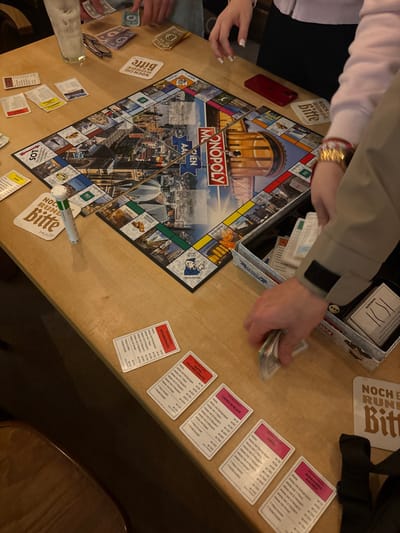 The image displays an overhead view of a Monopoly board game in progress on a light-colored wooden table. The game board is a specialized "MONOPOLY AACHEN" edition, indicating the location is the city of Aachen, Germany. At least three individuals are participating, evidenced by visible hands: one pair (top-left) is handling Monopoly banknotes, another hand (lower-right, with a beige jacket sleeve) is sorting a stack of Monopoly money, and a third hand (mid-right, with multiple gold-colored bracelets) is reaching into the game's white cardboard box to retrieve cards.
On the table, several Monopoly property cards from the Aachen edition are laid out face-up, including "KLEINMARIASTRASSE" (red), "ZENTIS JÜLICHES EDELLÄDCHEN" (red), "LADIES IN BLACK" (pink), "TIVOLI" (pink), and "ALEXANDRA AACHEN" (pink). Scattered Monopoly money in various denominations (e.g., green, purple, yellow) is present. A clear glass containing a beverage and ice cubes, a pair of silver-rimmed reading glasses, and a small white cylindrical container with a green cap are positioned on the left side of the table. A red smartphone rests on the table near the board's right edge. Two beige square coasters featuring the German phrase "NOCH EINE RUNDE BITTE" are also visible. The environment suggests an indoor setting, likely a commercial establishment such as a bar or cafe, under moderate lighting.