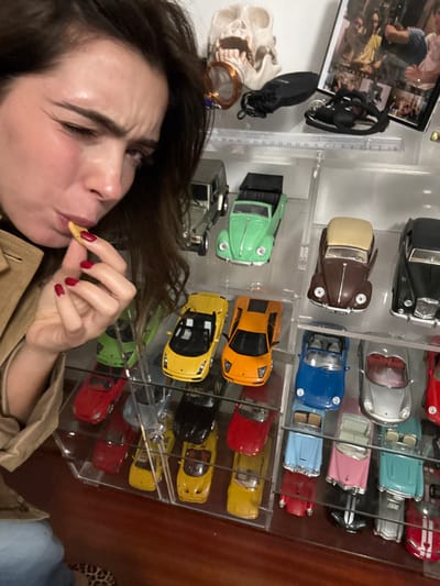 A woman with dark hair and red nails is eating a snack in front of a clear cabinet filled with miniature cars. The cars are neatly arranged in rows and feature various colors and models. A white skull, black sneakers, and a framed photograph are visible to the left of the cabinet. The woman is wearing a tan jacket.