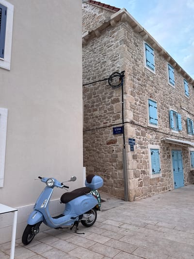 A light blue Vespa scooter is parked on a paved sidewalk in front of a stone building with blue shutters. The building is located on a narrow street in Primošten, Croatia. The street is paved with light gray stones. There is a white building on the left side of the image. The sky is cloudy.
