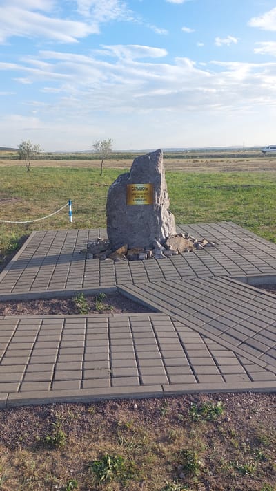 A large boulder, possibly a monument, stands on a paved square in Zhartas, Kazakhstan. The boulder has a golden plaque with writing on it. The paved area is made of grey interlocking bricks and is surrounded by a strip of dirt and grass. Two small, bare-branched trees are visible in the background, along with a grassy field and a distant horizon under a cloudy blue sky. A white van is partially visible on a road in the far distance.