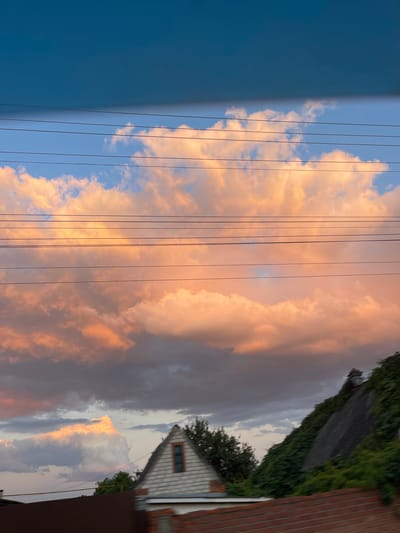 The image captures a sky with large, fluffy clouds illuminated with warm, peachy-orange hues, suggesting either sunrise or sunset. Several horizontal power lines crisscross the sky, dividing the frame. Below the sky, the upper part of a house with a peaked roof and a small window is visible. To the right of the house, a steep, dark slope is densely covered in green foliage, with a chimney visible at its peak. In the foreground, parts of a brown roof and a brick structure are partially obscured, indicating a residential area. The scene is set in the city of Krasnokutsk, Ukraine.