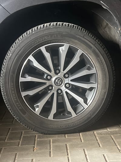 The image shows a close-up of a car wheel, specifically a tire and rim. The tire is a Dunlop Grandtrek AT30 with the size designation 265/55R19 109V visible. The rim is a multi-spoke alloy design with silver and black accents. The Toyota logo is present in the center of the rim. The car appears to be parked on a paved surface, possibly a driveway or parking lot, composed of grey brick pavers. The background is dark and out of focus, suggesting the car is in a shaded area or under artificial light. There is no indication of people or any action taking place in the image. The context of Dubingiai, Lithuania, is not visually represented in the image itself.