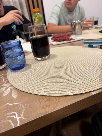 The image captures a casual indoor scene around a table in Dolno Sahrane, Bulgaria. The photograph is tilted clockwise, showing a close-up perspective.
**Subjects:**
Two individuals are partially visible. In the upper left quadrant, a person, likely male, wearing a light-colored striped shirt, is holding a clear glass. Their face is out of focus. In the lower left, the arms and upper torso of another person, wearing a black t-shirt, are visible, appearing to be handling a small, light-blue object.
The scene centers on a light-colored wooden table, partially covered by a light brown tablecloth adorned with a subtle, lighter-toned floral or leaf pattern. A large, round, light beige woven placemat with a distinct spiral pattern occupies a significant portion of the image.
Several glasses are present: a clear glass filled with a dark beverage (resembling cola) resting on the placemat, a decorative blue embossed glass, and another clear glass partially visible in the lower left.
A white rectangular plate holding sliced reddish cured meat (possibly sausage or salami) is placed near the person in the upper left. A plastic bottle with an orange and green label, likely juice, is partly visible in the lower left. Additionally, a slender, blue-metallic object, possibly a vape pen, lies near the top edge of the placemat.
**Activity or event taking place:**
The arrangement of food, drinks, and people suggests an informal meal, snack time, or gathering.
**Notable details:**
The image is captured under artificial indoor lighting. The general atmosphere appears relaxed and casual. The specific location of Dolno Sahrane, Bulgaria, is provided contextually.
**Visible text:**
Text partially visible on the orange and green bottle includes "Vita..." along with other indistinct words. A barcode and unreadable small text are present on the blue-metallic object.