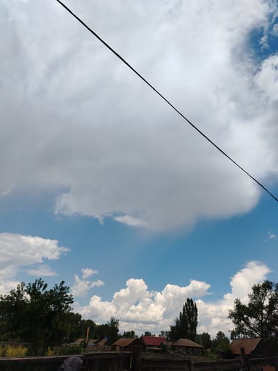 This media file captures a rural landscape under a partly cloudy sky. Dominating the upper portion of the image is a thick, wispy cloud formation casting a soft light over the scene. A dark, diagonal line, likely a power cable or wire, stretches across the sky, dividing the frame. Below this, a clear blue sky peeks through, dotted with smaller, fluffy white clouds.
The lower half of the image reveals a village or hamlet. Several houses with traditional, sloped roofs are visible, some appearing to be made of wood and others with corrugated metal roofing. A dense line of trees, some dark and leafy, others taller and more slender, forms a backdrop to the settlement. A wooden fence runs across the foreground, partially obscuring the view of the houses and vegetation behind it. A dark object, possibly a piece of clothing or a bag, rests on the fence. The overall impression is one of a tranquil, everyday scene in a rural setting. The time of day appears to be daytime, likely late morning or early afternoon, given the bright sky and the way light falls on the clouds and landscape. The weather seems fair, with the clouds suggesting a possibility of light rain or simply a dynamic sky. The location is identified as Zhovten village in Kazakhstan. There is no visible text in the image.
