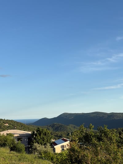 The provided image, when viewed in its correct landscape orientation, depicts a panoramic view of a mountainous coastal landscape under a clear blue sky. The upper two-thirds of the frame are dominated by a bright blue sky, gradually lightening towards the horizon, with sparse, wispy white clouds scattered across it. A distinct horizon line separates the sky from a vast body of water, likely the Adriatic Sea, which appears a pale blue due to distance and atmospheric haze.
Below the water, a range of dark green, densely forested mountains and hills stretches across the middle ground, their peaks forming an undulating silhouette against the lighter horizon. In the lower right foreground, a sloping hillside is covered with lush green vegetation, including various trees and bushes. Two buildings are visible on this slope: an upper, light-colored house with a reddish roof, featuring what appear to be solar panels and an exterior air conditioning unit, and a larger, more modern light-grey building with dark windows situated further down the hillside. A paved road or path can be seen winding near the lower structure.
The scene portrays a bright, sunny day with clear weather. No people or specific activities are visible, presenting a tranquil, natural landscape combined with some residential development. Given the context, the location is Pelinovo, Montenegro, which aligns with the visual characteristics of a mountainous Mediterranean coast. No discernible text is present in the image.