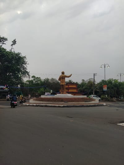 The image shows a roundabout in Chopdem, India, featuring a bronze-colored statue of a man with his right arm raised. Behind the statue, a partially visible building with red and yellow decoration can be seen. The roundabout is landscaped with reddish-brown soil and some foliage. Several motorcycles are on the road near the roundabout. Overhead utility lines and streetlights are also visible. The sky is overcast.