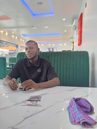 The image shows a man wearing a black shirt that says "jeen sale" sitting at a table in a restaurant in Ujevwu, Nigeria (latitude: 5.471, longitude: 5.818). He is looking at the camera and has his hands on the table. There is a green booth behind him, and a white table with a white marble top in front of him. A pink and blue checkered napkin is lying on the table. There is a sticker on the table that says "Scan me - Please pay to only AMARA LUXE HOTEL & SUITES ACCOUNT." The background shows a white ceiling with red and blue lights, and a window looking out onto an outdoor area. A TV screen on the wall shows an unidentified person. The restaurant appears to be clean and well-lit. There are other people in the background, but they are not visible. The image was taken at a medium distance and the camera is slightly angled upward.