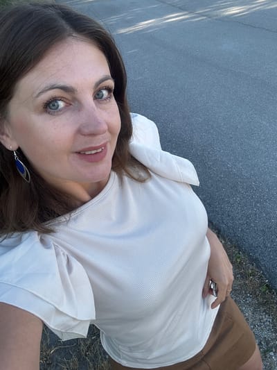 A woman with brown hair and blue eyes is taking a selfie outdoors. She is wearing a cream-colored top with puffy sleeves and a brown skirt. She has blue earrings with silver accents. She is standing on a paved surface, possibly a road or sidewalk, with some dry grass and gravel to the side. The background is out of focus, but appears to be a natural outdoor setting. The lighting suggests it is daytime. There is no indication of the specific location within Lavans-lès-Saint-Claude, France.