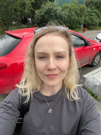 A fair-skinned woman with blonde, curly hair is taking a selfie outdoors. She is wearing a dark grey long-sleeved shirt and a silver necklace with a circular pendant. Her hair is partially covering her face. Behind her is a bright red car, a curb, and some greenery. The setting appears to be a parking area or street with trees in the background. The scene is captured in daylight.