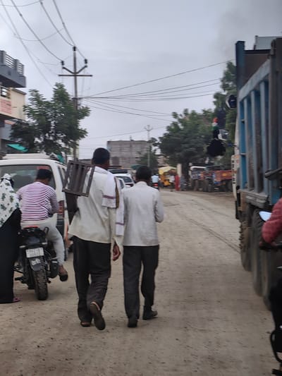 In Padli, India, on a cloudy day, two men walk away from the camera on a dirt road. The man on the left, wearing a light-colored shirt and dark pants, carries a metal rack on his back. The man on the right, also in dark pants, wears a light-colored shirt. A white van is parked to the left of the men, with a person wearing a striped shirt riding a motorcycle beside it. A large blue truck is on the right side of the road, partially obscuring the view of other vehicles and trees in the background. Electrical wires and a utility pole are visible in the sky.