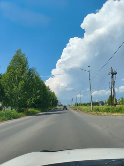 This is a photograph taken from a vehicle, looking forward along an asphalt road. On the left side of the road is a dense line of green trees, possibly birch trees, with a bright yellow sign visible amongst them. The right side of the road features a grassy verge with several utility poles and power lines. In the distance, several modern apartment buildings are visible, suggesting an urban or suburban setting. The sky is a bright blue, with a large, dramatic white cumulus cloud dominating the upper right portion of the frame. A single dark-colored car is driving away from the camera in the middle of the road. The overall impression is a sunny day with a sense of forward motion. This scene is consistent with locations in or around Металлплощадка, Russia, given the style of architecture and the presence of power infrastructure.