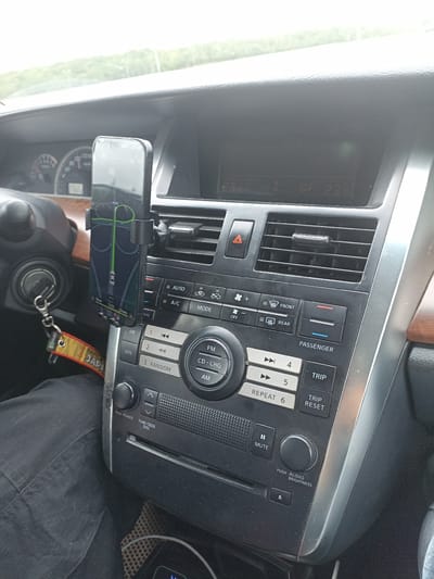 This media file is a close-up photograph taken inside a car, focusing on the central dashboard area. The image captures a smartphone mounted on a car mount, displaying a navigation app with a route highlighted in green. The phone screen is partially reflective, showing some smudges.
To the left of the phone, a portion of the steering wheel and ignition are visible. A keychain with a yellow and red tag featuring Japanese text ("MALAYSIA JEPANG" appears to be visible from the crops) hangs from the ignition. The car's dashboard features various controls, including buttons for climate control (AUTO, A/C, MODE), radio functions (FM, AM, CD-CHG), and trip information (TRIP, TRIP RESET). There are also several vents and a display screen showing digital readouts, seemingly indicating temperatures of "23.0" and "23.6". The interior of the car is dark, suggesting it might be daytime with overcast skies, as indicated by the bright, diffused light coming from outside, and the lack of harsh shadows. The overall scene is a snapshot of a car's interior, likely during a journey. No people are clearly visible in the frame, though the presence of a driver's leg covered in black fabric suggests someone is operating the vehicle. The car appears to be parked or stopped, given the focus on the dashboard and navigation system.