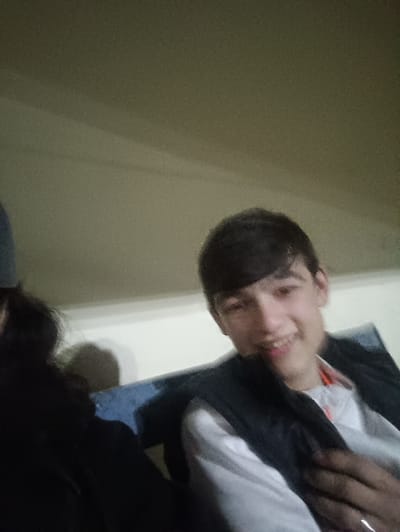 The image captures a close-up, informal portrait of two individuals, likely indoors. The person on the right is prominently featured, a young male with short, dark hair styled with a fringe. He is smiling broadly, showing his teeth, and looking directly at the camera. He is wearing a white long-sleeved shirt underneath a dark, possibly black or navy, sleeveless vest or jacket. His right hand is visible in the lower right corner, resting on his chest or arm, and he has a ring on one of his fingers, possibly the ring finger.
To the left, another person is partially visible. Only the left side of their head, with long, dark hair, and a portion of their upper body, dressed in a dark garment (possibly a hoodie or jacket), can be seen. Their face is entirely out of frame or obscured.
The background consists of a plain, light-colored wall, likely off-white or beige, with a slight texture visible. A darker, possibly blue, object, which appears to be part of a seat or a low barrier, is positioned behind the individuals. The lighting in the scene is somewhat dim, suggesting an indoor setting with either artificial or indirect light. The overall mood conveyed by the visible individual's smile is cheerful and relaxed. No text is visible in the image.