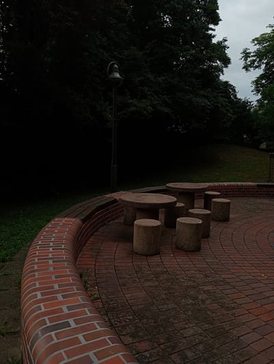 This image depicts an outdoor seating area with a stone table and stools, situated on a circular paved surface made of red bricks. A low, curved brick wall surrounds the seating area. In the background, a tall lamppost stands against a backdrop of dark green trees and a cloudy sky. The scene appears to be in a park or public space, likely in Constance, Germany, given the context. There are no people or animals present in the image, and no specific actions or interactions are occurring. The environment suggests a natural setting with urban amenities.