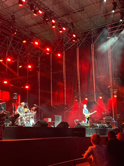 This image captures a live music performance on a stage at night. The stage is set up outdoors, under a metal scaffolding structure that holds numerous red lights, some of which are actively illuminating the stage. The background is dominated by dark red drapes.

On the stage, two musicians are visible. To the left, a male musician in a grey t-shirt and light-colored pants is playing a guitar and appears to be singing into a microphone. Behind him is a drum kit. To the right, another male musician, wearing a blue t-shirt and dark pants, is playing a yellow guitar. There are several black speakers placed on the stage, and some smoke or fog is present, catching the red light.

In the foreground, the backs of several audience members are visible, watching the performance. They are seated or standing behind a barrier. The overall lighting is intense and dominated by red hues.

This scene is located at the Bahía de Cádiz Auditorium in Chiclana de la Frontera, Spain, which is known for hosting concerts and other cultural events.