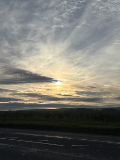 The image shows a rural landscape with a road in the foreground and a hazy sunrise in the background. The sky is filled with scattered clouds, and the sun is peeking through, casting a warm glow. The land appears to be agricultural, with fields of crops and rolling hills. The road is asphalt and appears wet, possibly from rain. There are no people or animals visible in the image. The scene is serene and peaceful, suggesting an early morning in the countryside.