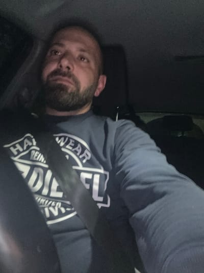 A male individual is depicted inside a vehicle, illuminated by an unseen light source in a dark interior environment. The individual, appearing to be an adult male with short or receding dark hair and a full dark beard, is positioned in the driver's seat or front passenger seat, with a black seatbelt diagonally across his torso. He is wearing a dark blue long-sleeved shirt featuring a white circular logo with text, partially visible as "HAR WEAR" at the top arc, "REIN" beneath, and "DIESEL" (or similar letters "DIE EL") at the bottom arc. His gaze is directed upwards and to his left, with a neutral to pensive expression. The background of the vehicle interior is indistinct due to low light conditions, revealing only dark surfaces and subtle outlines of car components. No identifiable landmarks or specific geographical features indicating the city of Hertsmere, United Kingdom, are visible within the image frame.