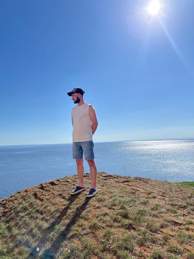 A man with a beard, wearing a white tank top, denim shorts, and a baseball cap, stands on the edge of a rocky cliff overlooking a body of water on a sunny day. The location is Шуран, Russia. The water glistens, and the sky is clear blue.