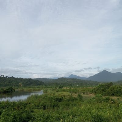The image shows a scenic landscape with a body of water in the foreground, surrounded by lush green vegetation. In the background, rolling hills covered in dense forest stretch towards the horizon, with mountains visible in the distance. The sky is overcast with a soft, diffused light. There are no people or animals visible in the image. The location appears to be rural, possibly in the vicinity of Babah Dua, Indonesia, given the tropical vegetation and mountainous terrain. The scene is tranquil and natural, depicting a serene natural environment.