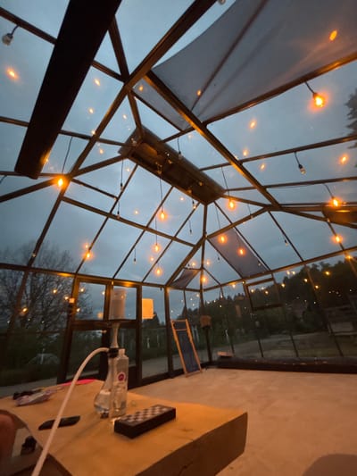 The image shows the interior of a greenhouse-like structure in Vlado Trichkov, Bulgaria. The structure has a glass and metal frame, with string lights illuminating the interior. Outside, a twilight sky is visible along with trees and some distant buildings.
Inside, a table occupies the right side of the frame. On the table are several objects including an electronic device (remote), and an item resembling a makeshift hookah made from plastic bottles.