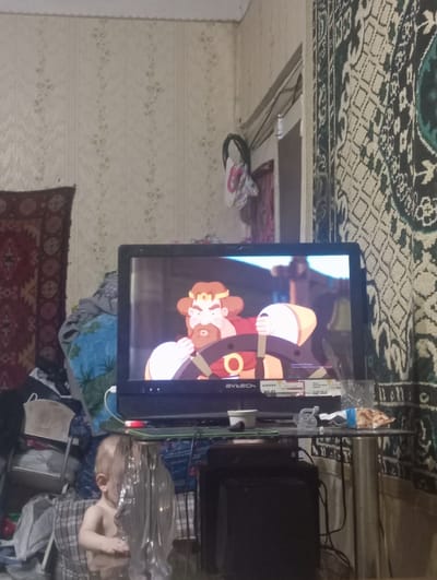 The image depicts an indoor scene, likely a living room, given the presence of a television, patterned carpet on the floor, and wallpaper. A television screen displays an animated character with orange hair and a crown, holding a weapon. To the right of the television, a portion of a black object, possibly another television or electronic device, is visible. Further to the right, a child, partially obscured by reflective material, is seated. The child appears to be looking towards the television. The room is cluttered with various items, including blankets and bags. The overall impression is one of a casual domestic setting. The time of day and weather conditions are not discernible. The location context provided is Nurabad, Uzbekistan. No visible text is clear enough to be read.