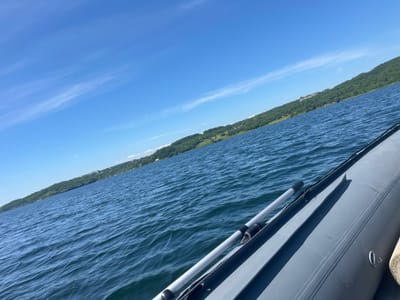 The image depicts a body of water, likely a lake or river, under a clear blue sky with scattered wispy clouds. The water is a deep blue with visible ripples, suggesting a gentle breeze. On the right side of the frame, the edge of a gray inflatable boat is visible, with an oar resting inside. The boat's tube runs along the bottom right and extends diagonally upwards. The distant shore is lined with lush green trees, indicating a natural, perhaps forested, landscape. A few structures, possibly buildings or houses, are visible nestled among the trees on the hillside. The overall scene suggests a calm, outdoor environment, likely during daylight hours. The provided hints indicate the image was taken from a boat looking towards the shore and the sky. The location of Zavety Il'icha, Russia is not visually identifiable from the image itself.
