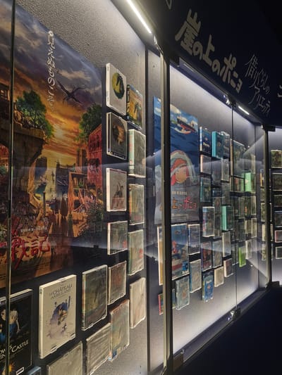 The image displays a museum exhibit likely at the Ghibli Park in Nagakute, Japan, showcasing Studio Ghibli films. The scene is dimly lit, with spotlights illuminating display cases filled with various film cases and posters. A large poster of "Ponyo on the Cliff by the Sea" is prominently featured, along with other artwork and film titles such as "Howl's Moving Castle" and "Le Château Ambulant." The walls are textured and dark, with visible lighting fixtures. The exhibit appears to be behind glass, suggesting it is a protected display. There are no people visible in the frame. The overall impression is one of a curated collection of animated film memorabilia.
