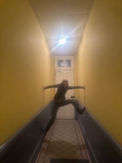 The image displays an indoor hallway in Roanne, France. A single adult individual is captured in an athletic pose, suspended horizontally across the width of the corridor. The person is smiling at the camera, wearing a dark jacket, dark pants, and a patterned cap. Both arms are extended, palms pressed against the light yellow upper walls for support. The legs are spread wide, with the right foot (viewer's perspective) braced against the dark blue lower wall section and the left foot higher against the yellow wall. The hallway features light yellow painted upper walls and a dark blue lower dado section. The floor is covered with geometric pattern tiles in shades of grey and white, with a dark mat visible at the foreground. At the far end of the hallway, a white door with two small, frosted glass windowpanes is visible. The ceiling is composed of a grid of square acoustic panels. A single, bright overhead light fixture centrally illuminates the scene.